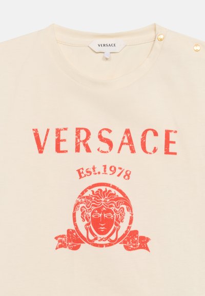 Κρεμ T-shirt με κόκκινο λογότυπο Versace, "Est. 1978," και γραφικά με κεφάλι της Μέδουσας, με δύο χρυσά κουμπιά στον ώμο.
