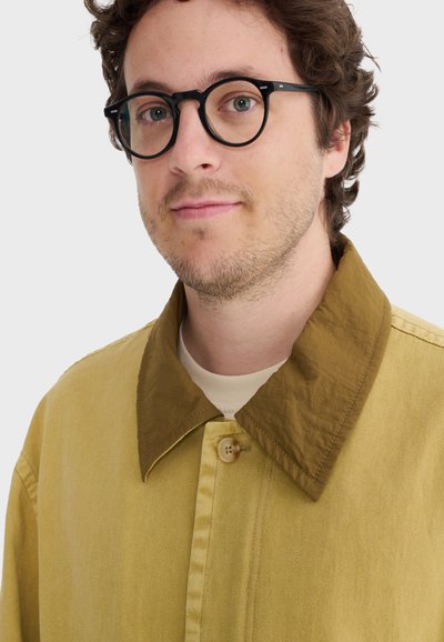 Uomo con capelli ricci che indossa occhiali rotondi neri e una giacca beige con un ampio colletto marrone su uno sfondo semplice.