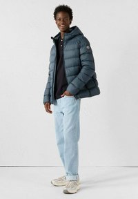 Chaqueta acolchada azul oscuro con capucha, combinada con una camiseta polo negra y vaqueros azul claro. Calzado: zapatillas beige con detalles texturizados.