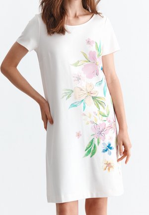 Kobieta ubrana w białą sukienkę z krótkim rękawem, zdobioną dużym pastelowym kwiatowym wzorem po jednej stronie, stojąca z jedną ręką na biodrze.