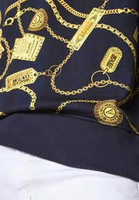 Tissu bleu marine avec motif chaîne dorée et étiquette affichant les textes « Versace Jeans Couture » et « Milano », associé à un pantalon blanc.
