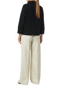 Giacca nera a tessuto strutturato con colletto alto, tasche laterali e maniche a tre quarti, abbinata a pantaloni a gamba larga color crema.