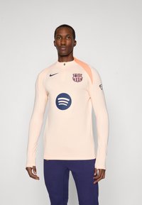 Man wearing a light pink FC Barcelona zip-up sports top with Spotify logo and navy blue pants, standing against a plain background.