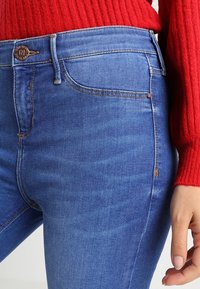 Blå denimjeans med slim fit. Funktioner inkluderar en synlig midjeband, fickor och kontrasterande sömmar. Tyget har en mjuk textur.