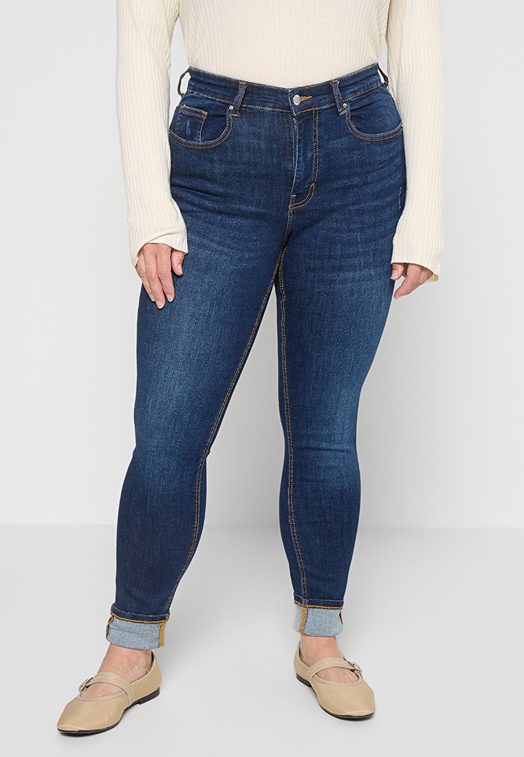Dunkelblaue, hochtaillierte Jeans mit heller Naht, zwei Fronttaschen und umgeschlagenen Säumen. Kombiniert mit beigen Ballerinas und einem hellen gerippten Oberteil.