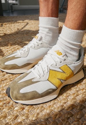 Person iført hvide og beige New Balance sneakers med gul "N"-logo og hvide sokker, stående på et vævet tæppe af naturlige fibre.
