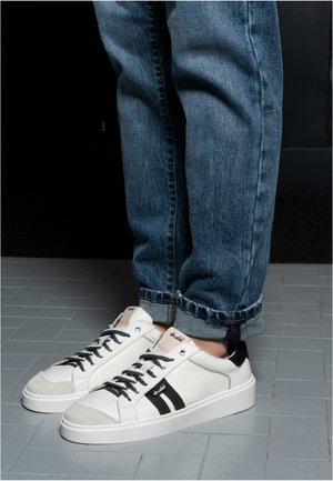 White low-top sneakers with black stripes and laces worn with rolled-up blue jeans on gray tiled floor.