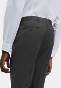 Mørkegrå skræddersyede bukser med en glat overflade, udstyret med en baglomme med knaplukning og båret med en hvid skjorte.