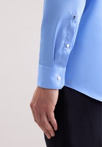 Camicetta azzurro chiaro con una texture liscia, polsini abbottonati e un logo a forma esagonale vicino alla spalla. Il tessuto appare elegante e su misura.