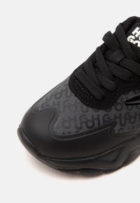 Gros plan sur la pointe d'une basket noire avec une tige à motif, des lacets noirs épais et une semelle texturée sur fond blanc.