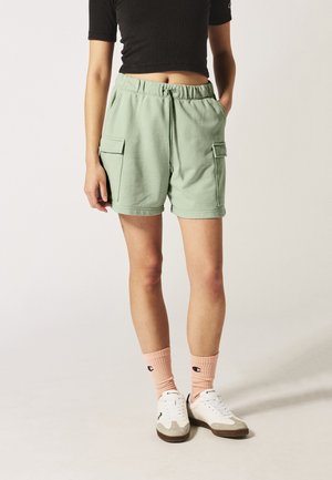Lichtgroene shorts met een elastische tailleband, twee zijzakken en een trekkoord. Gecombineerd met witte sneakers en roze geribbelde sokken.