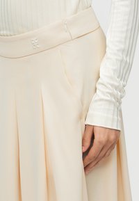 Μπεζ φούστα με πιέτες και πλαϊνό φερμουάρ, φορεμένη με μακρυμάνικο ριμπ εκρού τοπ, που δείχνει κοντινό στην μέση και το χέρι.