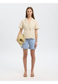 Blazer beige à rayures, à manches courtes, double boutonnage, associé à un short en denim clair. Le modèle tient un sac tissé texturé.