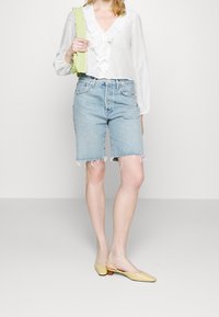 Blouse blanche à volants, short en denim bleu clair avec ourlet effiloché, et chaussures à bride arrière jaunes. Le modèle tient un sac vert citron.