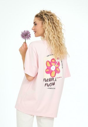 Kort, lyserød, oversized t-shirt med et farverigt blomstertryk og tekst på ryggen. Model der holder en lilla blomst, set fra siden.