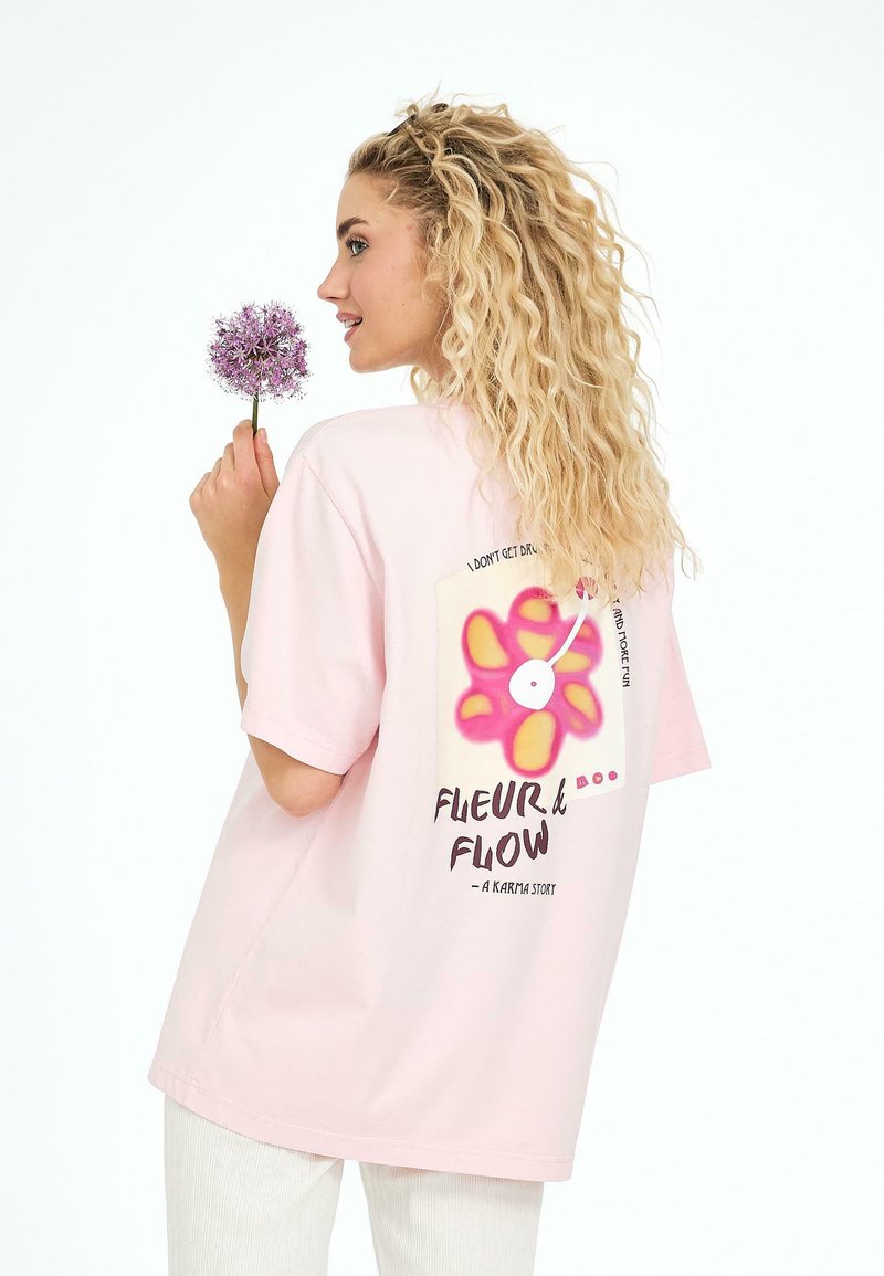 T-shirt surdimensionné rose, avec un graphique de fleur coloré et du texte au dos. Modèle tenant une fleur violette, vu de côté.