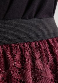 Ceinture élastique noire sur une jupe en dentelle bordeaux avec des motifs floraux et un tissu texturé, mettant en valeur un design superposé et un savoir-faire détaillé.