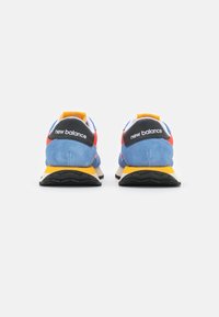 Siniset mokkakengät mustilla yksityiskohdilla, keltaisella pohjalla ja punaisilla yksityiskohdilla. "New Balance" -logo näkyvästi kantapäässä.