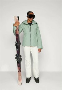 Skijacke in hellgrün, mit Reißverschluss und Kapuze; kombiniert mit weißen Skihosen und schwarzen Skischuhen; hält einen gemusterten Ski mit Bindungen.