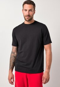 Hombre con el brazo izquierdo tatuado, vistiendo una camiseta negra sencilla y pantalones rojos, de pie contra un fondo claro y liso.