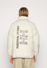 Pagrįsta smėlio spalvos striukė su aukštu apykakle. Ant nugaros yra tamsiai pilkas "Helly Hansen" logotipas. Elastingos rankogaliai ir apatinė dalis, kad puikiai laikytųsi.