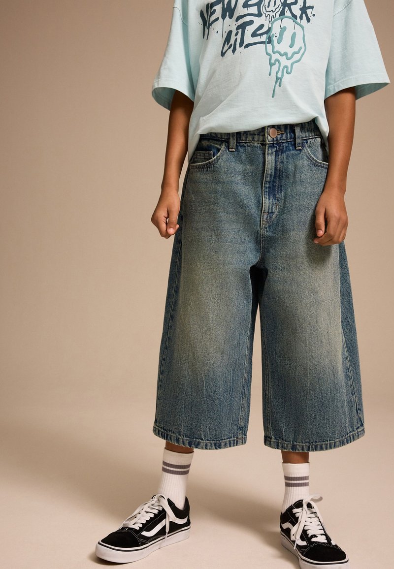 Culottes en denim, bleu clair avec texture délavée, taille haute, poches latérales. Associées à un t-shirt graphique et des baskets noires, chaussettes blanches.
