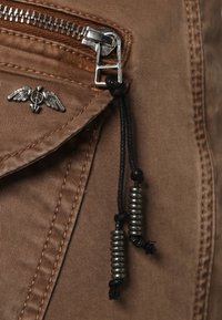 Brown fabric with a front zip pocket. Features a silver winged peace symbol and a string pull adorned with metallic coils.