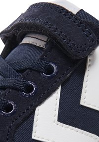 Mörkblå sneakers med mocka och textil, i kombination med vita läderdetaljer och blå öglor. Inkluderar snören och ett spänne med kardborre.