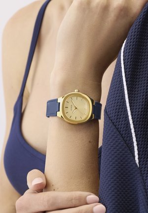 Montre-bracelet en or avec un cadran rond, un bracelet en silicone bleu et des index horaires minimalistes. Détail de la marque visible sur le cadran.