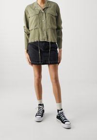 Chaqueta ligera de color verde oliva, abotonada, con dos bolsillos en el pecho, combinada con una falda de mezclilla negra y zapatillas altas negras.