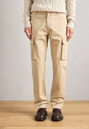 Pantalons cargo beiges en coton, dotés de poches latérales et d'une coupe décontractée. Associés à des chaussures marron foncé sur un sol en bois.