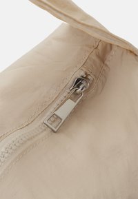 Prato in tessuto beige chiaro in primo piano, caratterizzato da una cerniera argento, cuciture intricate e una texture morbida, leggermente stropicciata.