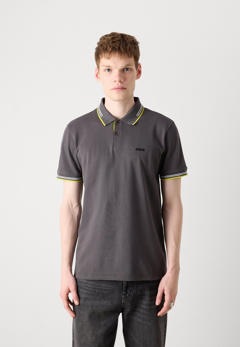 Graues Poloshirt mit einem Zwei-Knopf-Verschluss, einem Kragen mit weißen und gelben Streifen sowie einem kleinen schwarzen Logo auf der linken Brust. Kombiniert mit dunklen Jeans.