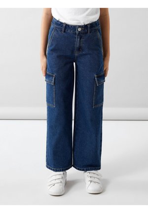 Enfant portant un pantalon cargo en denim bleu à jambes larges avec poches à rabats et des baskets blanches à scratch, debout sur un sol clair.