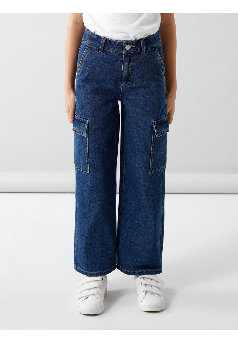 Enfant portant un pantalon cargo en denim bleu à jambes larges avec poches à rabats et des baskets blanches à scratch, debout sur un sol clair.