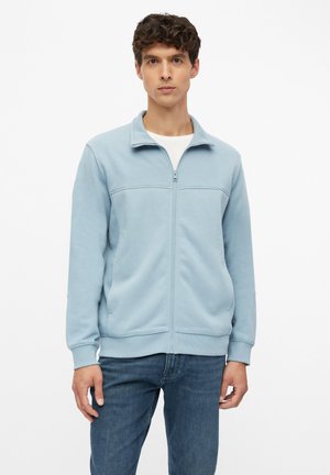 Mann trägt hellblaue Jacke mit Reißverschluss über weißem Hemd und blauer Jeans, steht vor einfachem weißem Hintergrund.