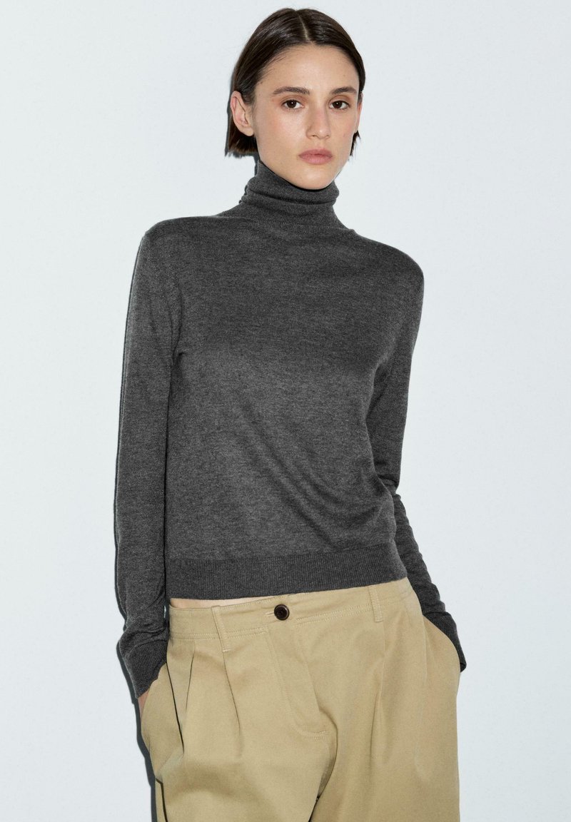 Giovane donna con capelli corti e scuri che indossa un maglione dolcevita grigio scuro e pantaloni beige a vita alta, in piedi con le mani nelle tasche.