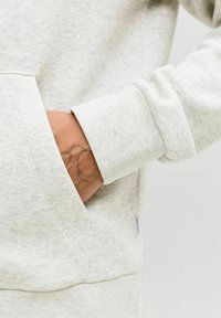 Sweatshirt gris clair avec une poche frontale, des manches avec poignets côtelés, et un tissu doux et texturé. Un tatouage est visible sur le poignet.