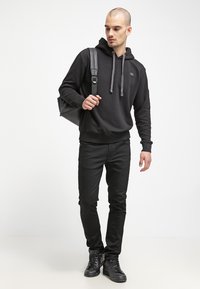 Sudadera negra con cordones, combinada con unos vaqueros negros ajustados y zapatillas negras. El modelo lleva una mochila oscura y texturizada.