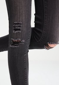 Jeans negros desgastados con desgastes deshilachados en las rodillas y los muslos. La tela parece elástica y ajustada, con un ligero desgastado.