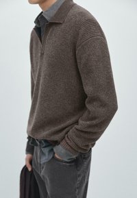 Pull en maille côtelée marron avec un col, porté sur une chemise en denim. La texture est épaisse et la coupe est décontractée avec des manches longues.