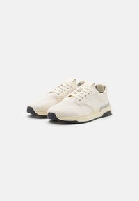 Beige knit sneakers with a textured upper, lace-up closure, light grey accents, and a black rubber outsole. Rounded toe design.