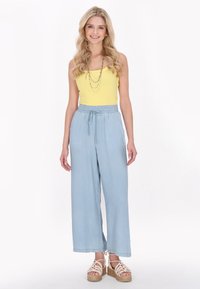 Geel geribbelde tanktop gecombineerd met lichtblauwe wijdpijpenbroek, met een verstelbare taille. Model draagt beige platform sandalen.