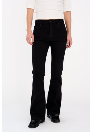 Kvinde iført sorte højttaljede flare-jeans, hvid cropped top og sorte loafers, stående mod en hvid baggrund.
