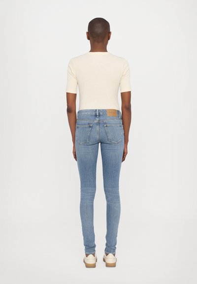 Jeans ajustados azules con una cintura de media altura, que presentan un parche de cuero marrón en la parte posterior y dos bolsillos traseros. Tela de denim ligero.