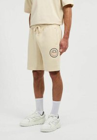 Bermudas de algodón beige con una cinturilla elástica, cordón y un logo impreso circular en el lado izquierdo. Combinadas con zapatillas blancas y calcetines.