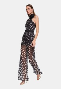 Tuta a pois bianco e nero con scollo all'americana, pantaloni larghi trasparenti e un dettaglio floreale sulla spalla. Sandali dorati in evidenza.