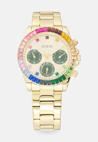 Gullfarget klokke med en bezel prydet med krystaller, grønn urskive, tre sub-dialer og et armbånd i rustfritt stål. Flersfargede steiner forbedrer designet.
