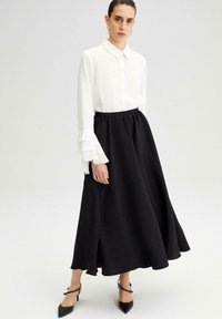 Bílá košile s knoflíky a volánkovými manžetami v kombinaci s volnou černou maxi sukní. Černé podpatky dotvářejí outfit. Hladká textilie.