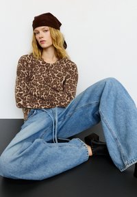 Sweter w panterkę w ciepłym brązie, połączony z luźnymi, jasnoniebieskimi jeansami. Całość dopełniają czarne botki na grubej obcasie i brązowa chusta na głowie.